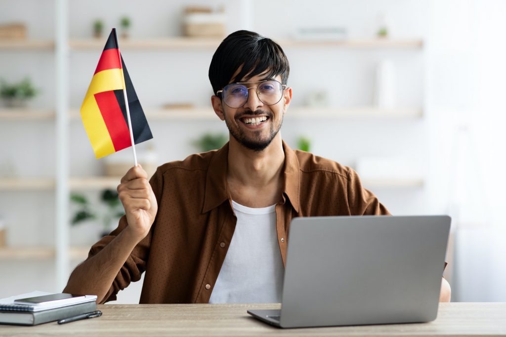Smiling indian guy student showing flag of Germany, using laptop